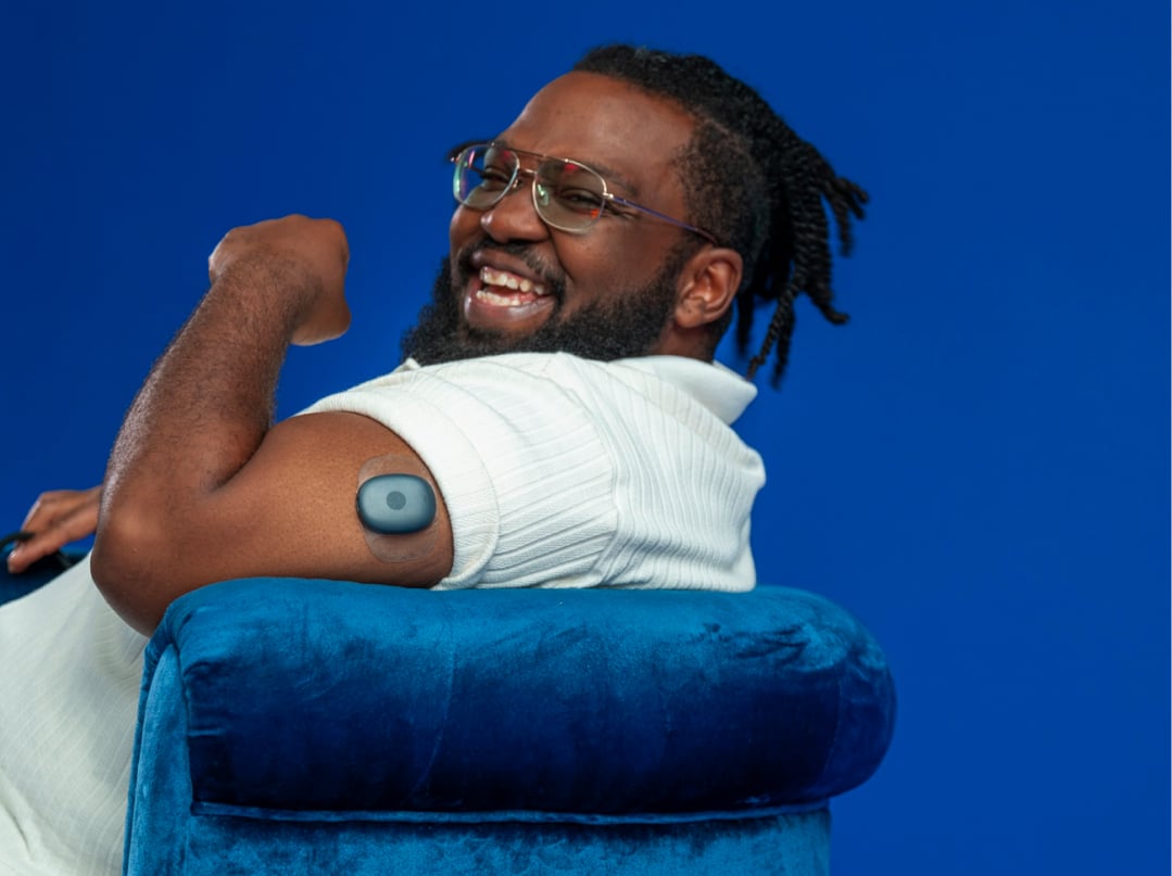 50 | 50 image Eversense user smiling, flexing arm with a CGM sensor device attached, seated on a blue chair.